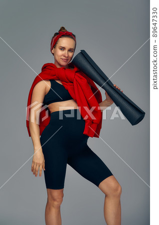 Pregnant woman holding yoga mat in hands, going for sport while awaiting baby, looking smiling at camera and touching her belly, standing against gray wall. 89647330