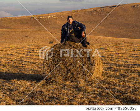 A man sits on a haystack in a wide golden field that is located in the Caucasian highlands on an autumn day. 89650055
