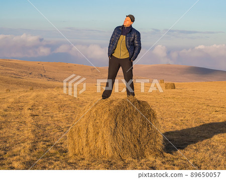 A man admires the picturesque surroundings while standing on a haystack in a wide golden field located in the Caucasian highlands. 89650057