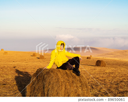 A woman in a bright yellow warm jacket sits in an autumn field on a haystack illuminated by bright sunlight 89650061