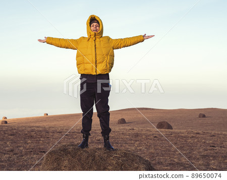 A cheerful rural girl stands with her hands apart on a haystack in a highland field under the sky 89650074