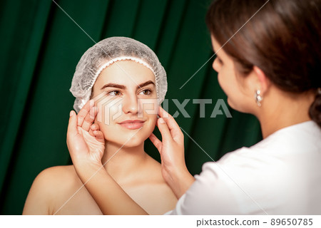 Plastic surgery and beauty concept. Professional cosmetician examining and touching the face of the beautiful young caucasian smiling woman in a clinic of esthetic cosmetology Plastic surgery and beauty concept. Professional cosmetician examining and touching the face of the beautiful young caucasian smiling woman in a clinic of esthetic cosmetology 89650785