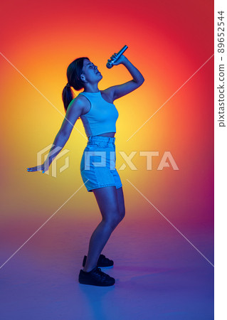 Emotive young girl singing in microphone, performing isolated over multicolored background in neon light 89652544