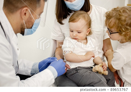 Male doctor stick a band-aid on a shoulder. Man wearing white medical gown and blue gloves. Boy wearing white plaid t-shirt. 89653767