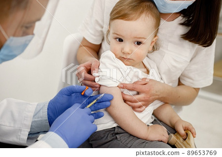 Mother holding her son on a lap. Medical worker wearing blue gloves and face mask. Boy wearing white t-shirt. Mother holding her son on a lap. Medical worker wearing blue gloves and face mask. Boy wearing white t-shirt. 89653769
