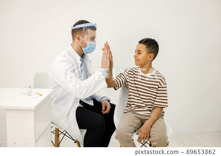 Male doctor sitting at table and wearing face mask. Man wearing white medical gown while isolated on white backgroun. Man give a high five to a little boy. Male doctor sitting at table and wearing face mask. Man wearing white medical gown while isolated on white backgroun. Man give a high five to a little boy. 89653862
