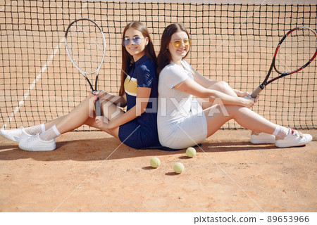 Two girls sitting on a tennis court and holding a rackets. Women wearing white and blue sport dresses and sunglasses. Girls standing together and posing for a photo. 89653966