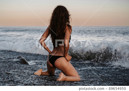 brunette girl with a sexy booty in a swimsuit is kneeling in the water in the waves of the sea 89654950