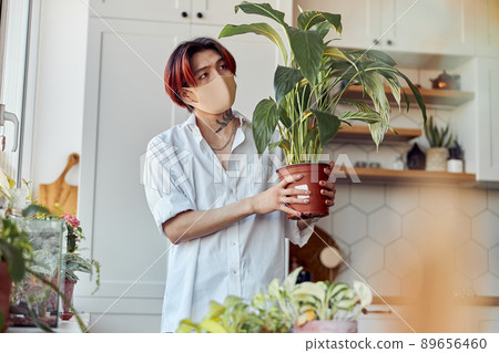 Young Asian man in mask holding flower pot with green plant near the window 89656460
