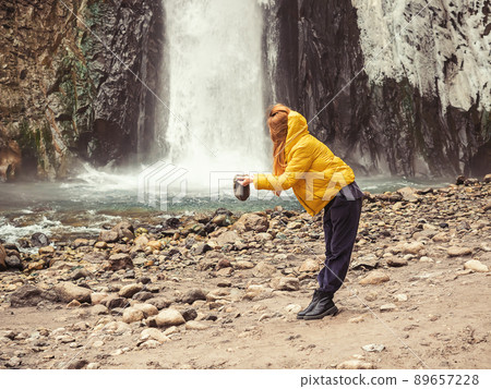 A woman stands against the backdrop of a stormy waterfall flowing down from wet, icy rocks, filling her hat with water from it. Optical illusion 89657228