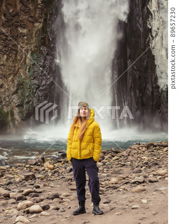 A woman stands on the river bank against the backdrop of a stormy waterfall flowing down from the rocks on a cold autumn day. Soft focus. A woman stands on the river bank against the backdrop of a stormy waterfall flowing down from the rocks on a cold autumn day. Soft focus. 89657230