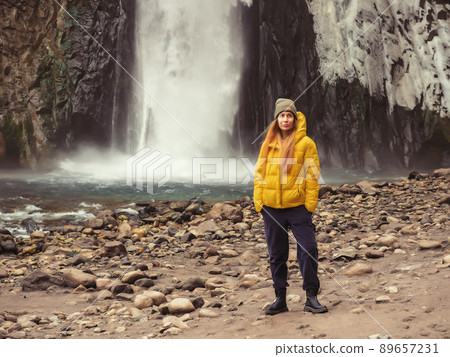The girl stands thinking against the backdrop of a large powerful waterfall flowing from an icy rock into a mountain river. Soft focus. 89657231