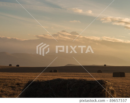 A wide field and haystacks against the backdrop of a mountain valley dissolving in a cloudy haze and the peaks of Elbrus visible above the clouds during sunset. Soft focus. A wide field and haystacks against the backdrop of a mountain valley dissolving in a cloudy haze and the peaks of Elbrus visible above the clouds during sunset. Soft focus. 89657337