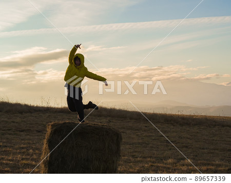 A woman in a yellow jacket in a dancing pose stands on one leg on a haystack in an autumn field high in the mountains at cloud level A woman in a yellow jacket in a dancing pose stands on one leg on a haystack in an autumn field high in the mountains at cloud level 89657339