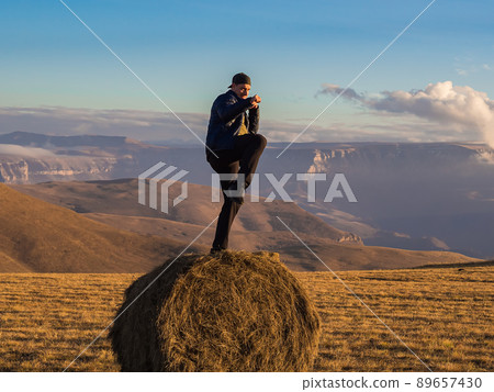A man stands in a fighting pose on one leg on a haystack in an autumn field which is located on the top surrounded by magnificent Caucasian mountains. A man stands in a fighting pose on one leg on a haystack in an autumn field which is located on the top surrounded by magnificent Caucasian mountains. 89657430