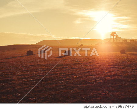 A golden field with haystacks on a mountain peak with a gazebo and tourists in the distance in a twilight haze and in the rays of the setting sun. Soft focus. A golden field with haystacks on a mountain peak with a gazebo and tourists in the distance in a twilight haze and in the rays of the setting sun. Soft focus. 89657479