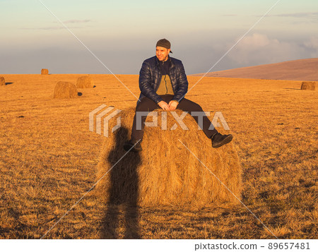 A smiling man lit by bright sunbeams sits on a haystack in an autumn golden field. A smiling man lit by bright sunbeams sits on a haystack in an autumn golden field. 89657481