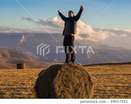 A man stands with his hands clenched into fists on a haystack in an autumn field which is located on the top surrounded by magnificent Caucasian mountains covered with clouds. 89657482