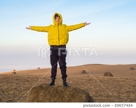 A girl in a yellow jacket stands with her hands apart on a haystack in a highland field under a blue sky A girl in a yellow jacket stands with her hands apart on a haystack in a highland field under a blue sky 89657484