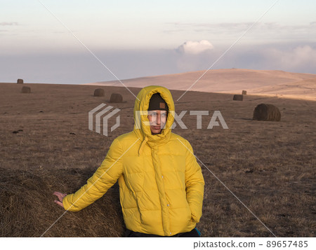 A girl in a yellow jacket, leaning on a haystack, stands in a wide field of the Caucasian highlands under a twilight sky A girl in a yellow jacket, leaning on a haystack, stands in a wide field of the Caucasian highlands under a twilight sky 89657485