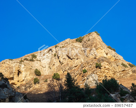 Illuminated by the bright sun, a sharp rocky mountain with coniferous green trees growing on it against the background of a blue sky Illuminated by the bright sun, a sharp rocky mountain with coniferous green trees growing on it against the background of a blue sky 89657487