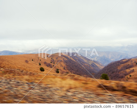 Autumn alpine valley covered with golden dry grass under low clouds. View from a passing car. Photo in motion 89657615