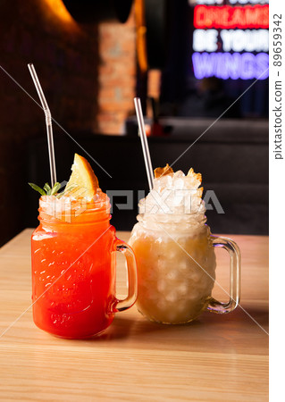Red Strawberry lemonade with soda, ice, cocktail tube and orange slice in glass with handle. Cocktail Pina Colada with ice, cocktail tube and pineapple chips in glass with handle on wooden table. 89659342