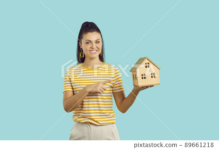 Smiling woman points finger at small wooden house mockup in her hand on turquoise background. Smiling woman points finger at small wooden house mockup in her hand on turquoise background. 89661218
