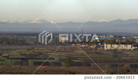Beautiful quiet spring panorama of plowed and green fields and meadows on background of trees, forest and distant view of magnificent mountain range with bright white snow shining under morning sun. 89663913