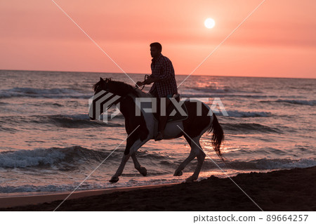 A modern man in summer clothes enjoys riding a horse on a beautiful sandy beach at sunset. Selective focus A modern man in summer clothes enjoys riding a horse on a beautiful sandy beach at sunset. Selective focus 89664257