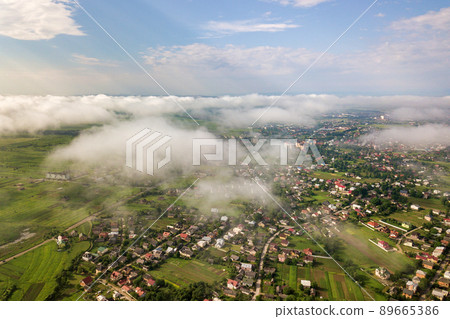Aerial view of white clouds above a town or village with rows of buildings and curvy streets between green fields in summer. Countryside landscape from above. Aerial view of white clouds above a town or village with rows of buildings and curvy streets between green fields in summer. Countryside landscape from above. 89665386
