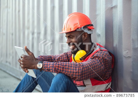 Close up happy smiling male African American engineer wearing safety vest and helmet, sitting and using mobile phone at logistic shipping cargo containers yard. Close up happy smiling male African American engineer wearing safety vest and helmet, sitting and using mobile phone at logistic shipping cargo containers yard. 89673785