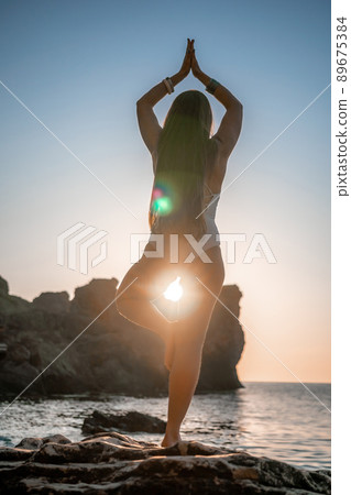 Young woman with long hair in white swimsuit and boho style braclets practicing outdoors on yoga mat by the sea on a sunset. Women's yoga fitness routine. Healthy lifestyle, harmony and meditation 89675384