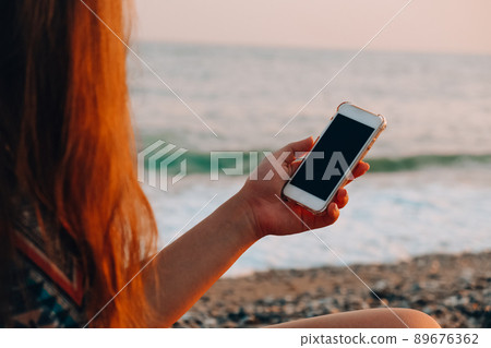 Unrecognizable young woman holding mobile phone at the beach against waves of sea. Sunset. Social media, modern technologies. Summertime vacations. 89676362