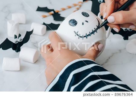 Female hands paint on white cup scary jacks face DIY for kids Halloween home activities Holiday art children craft Handmade decorations Step by step Socially distanced at home 89676364
