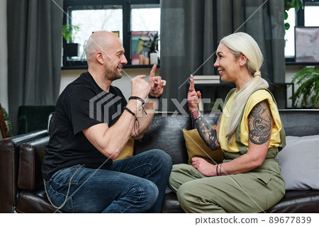 Side view shot of modern mature man and woman in love sitting on couch in living room communicating using sign language Side view shot of modern mature man and woman in love sitting on couch in living room communicating using sign language 89677839