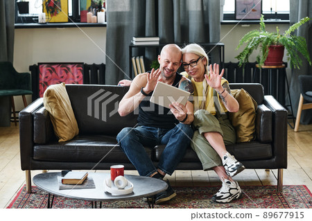 Stylish mature man and woman sitting on sofa in living room greeting their friends or relatives on video call on digital tablet 89677915