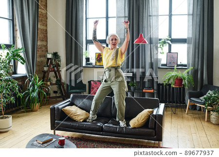 Stylish mature Caucasian woman with tattooes on arms listening to music in headphones and dancing on sofa in living room 89677937