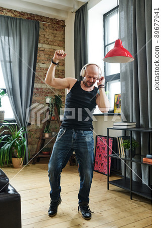 Vertical full body shot of mature Caucasian man wearing jeans and sleeveless shirt enjoying music in wireless headphones and dancing in living room Vertical full body shot of mature Caucasian man wearing jeans and sleeveless shirt enjoying music in wireless headphones and dancing in living room 89677941