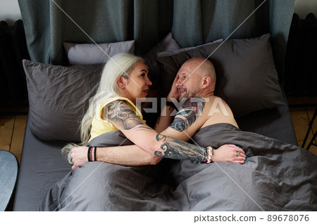 From above view of mature man and woman in live with tattooed arms relaxing in bed in morning talking about something From above view of mature man and woman in live with tattooed arms relaxing in bed in morning talking about something 89678076