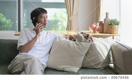 Relaxed asian man sitting on comfortable sofa at home and having conversation with mobile phone. Relaxed asian man sitting on comfortable sofa at home and having conversation with mobile phone. 89678486