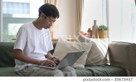 Man using laptop, sitting on couch at home, chatting or shopping in internet, reading message, working online with device. Man using laptop, sitting on couch at home, chatting or shopping in internet, reading message, working online with device. 89678487