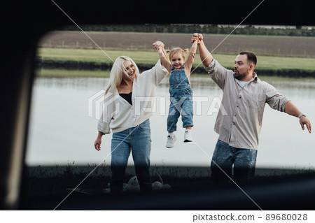 Happy Young Family Having Fun Outdoors, Mom and Dad Taking Up Their Little Daughter by Hands, View Through Window Inside of the Car 89680028
