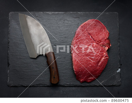 Raw beef tenderloin lies on a cutting board and spices for cooking on a black table, top view Raw beef tenderloin lies on a cutting board and spices for cooking on a black table, top view 89680475