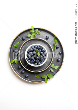 White bowl with washed wild blueberries and green leaves laid out on metallic plates on white. Blueberry - superfood rich in vitamins useful for health. Copy space. Top view. Vertical orientation. White bowl with washed wild blueberries and green leaves laid out on metallic plates on white. Blueberry - superfood rich in vitamins useful for health. Copy space. Top view. Vertical orientation. 89684127