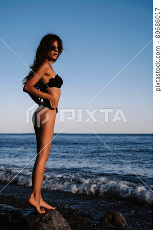 sexy girl brunette in a black swimsuit with a slender figure posing on the beach by the sea 89686017