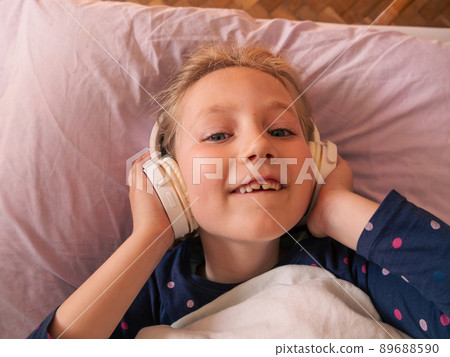 Blonde child girl in headphones learning language listening to music podcast with smartphone online in bed at home. Blonde child girl in headphones learning language listening to music podcast with smartphone online in bed at home. 89688590