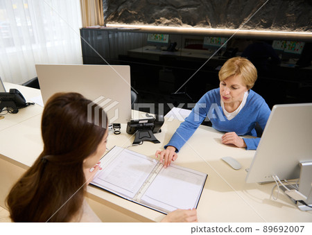 View from above to female sales manager, travel agent talking to her client, showing catalog with tours or consumer services, sitting in modern office 89692007
