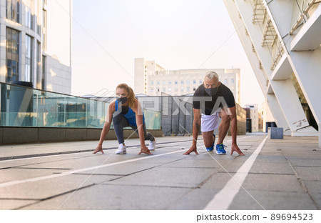 Sporty middle aged couple wearing protective masks on the start position ready for running outdoors during coronavirus outbreak Sporty middle aged couple wearing protective masks on the start position ready for running outdoors during coronavirus outbreak 89694523