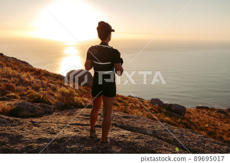 Woman in sportswear standing on the cliff and looking at the view at sunset. Female trail runner relaxing on mountain top looking at the ocean. Woman in sportswear standing on the cliff and looking at the view at sunset. Female trail runner relaxing on mountain top looking at the ocean. 89695017